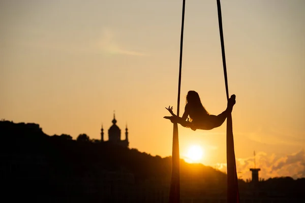 Silhouette of a flexible woman acrobat on aerial silk during a sunset on Kiev city background. concept of freedom and peace — Stock Photo, Image