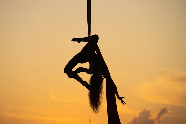 Silhueta de uma mulher flexível acrobata em seda aérea durante um pôr do sol. beleza conceito — Fotografia de Stock