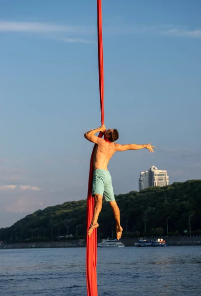 Izmos férfi cirkusz művész csinál őrült trükk légi selyem az ég és a folyó háttér — Stock Fotó