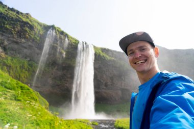 Arka planda Seljalandsfoss 'un güzel şelalesiyle selfie çeken genç adam. Seyahat ve sağlıklı yaşam tarzı kavramı