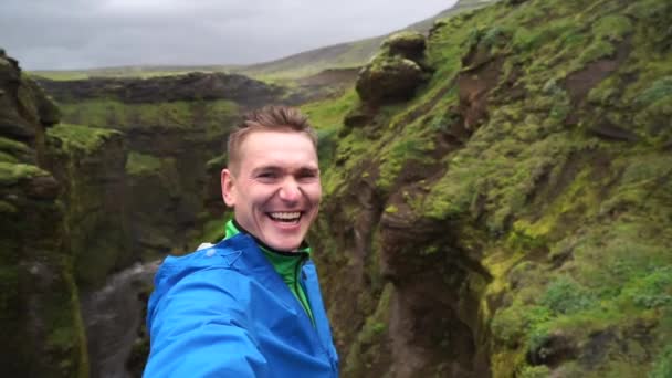 Молодой улыбающийся человек с рекой и зелеными горами на заднем плане делает селфи. Понятие свободы передвижения и свободы — стоковое видео