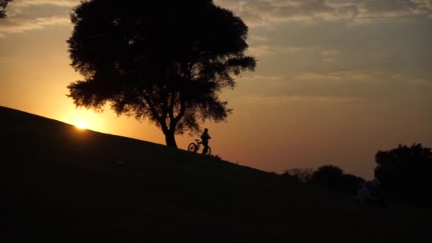 Silhouet van kind gaat bergafwaarts met de fiets bij zonsondergang in slow motion — Stockvideo