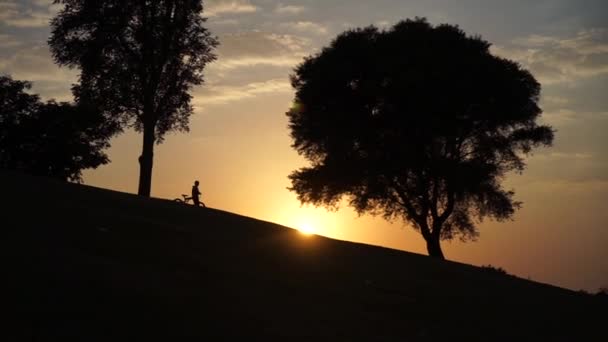 Silhouet van kind gaat bergafwaarts met de fiets bij zonsondergang in slow motion — Stockvideo