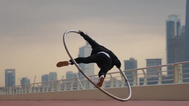 Художник Cyr Wheel делает трюки замедленного действия в черно-белой умной одежде на фоне города Дубая во время заката — стоковое видео