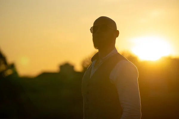 Bald Guy with a stylish beard and sunglasses on a blurred city background during dramatic sunset. Concept of success and will