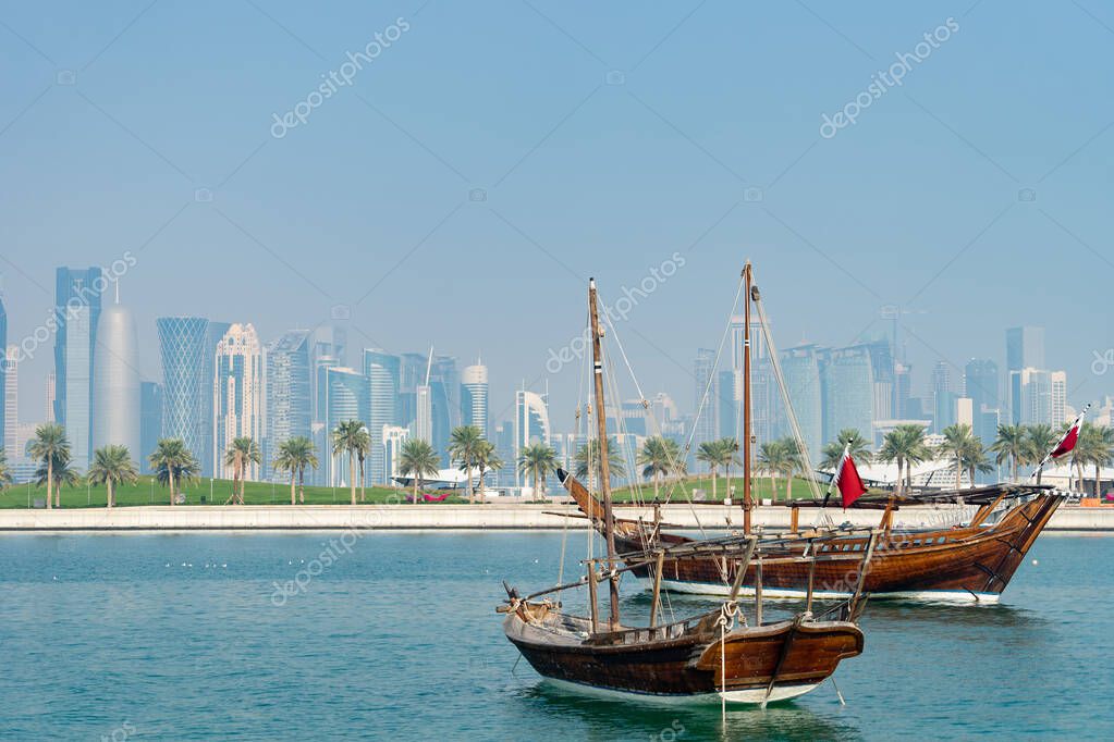 Retro historical boat with blurred panoramic view of modern skyline of Doha and green palms on background.