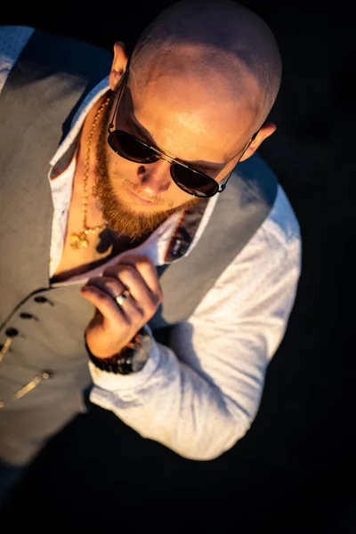 Bald Guy with a stylish beard and sunglasses on a blurred background during sunset. Concept of success and will