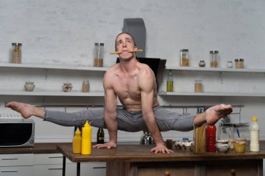 Kaslı adam mutfakta elleri dengede tutuyor ve ağzında makarna tutuyor. Mutfaktaki yaratıcılığı ve sağlıklı yaşam tarzını kavra.