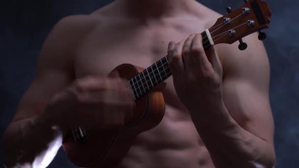 Jonge gespierde mannelijke muzikant speelt een ukelele ob zwarte achtergrond met rook — Stockvideo