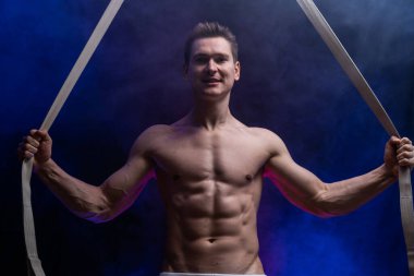 Siyah ve tütsülenmiş arka planda hava kayışları olan kaslı erkek sirk sanatçısı.