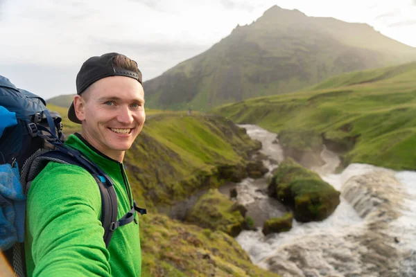 Joven hombre sonriente con río y montañas verdes en el fondo toma un selfie. Concepto de libertad de movimiento y libertad — Foto de Stock