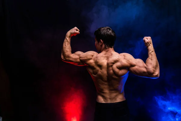 Hombre musculoso mostrando músculos aislados en el fondo negro con humo de color. Concepto de estilo de vida saludable — Foto de Stock