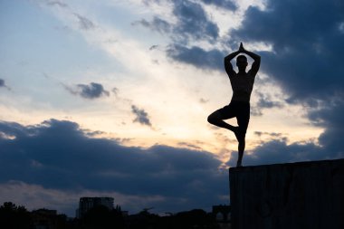 Formda genç adam gün batımında uçurumun kenarında güneş selamı yogası yapıyor.