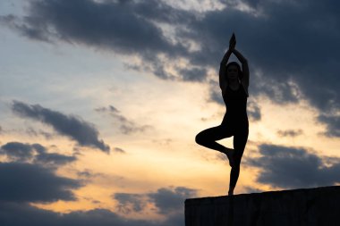 Formda genç kız gün batımında uçurumun kenarında güneş selamı yogası yapıyor.