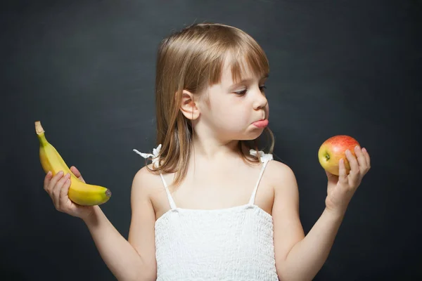 Little girl with apple and banan in the hands. The choice between fruits. Facial expression. — ストック写真