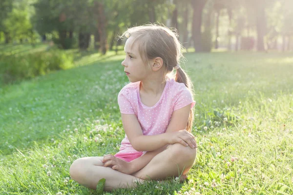Menina bonita em camiseta rosa na grama. Menina cansada após passeio no parque da cidade — Fotografia de Stock