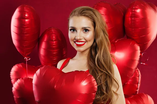 Menina atraente sorrindo e segurando balões de coração vermelho, retrato de beleza da moda — Fotografia de Stock