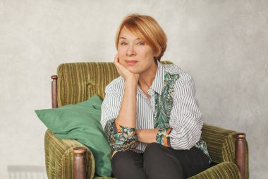 Evdeki yeşil koltukta oturan kısa kızıl saçlı, olgun bir kadın.