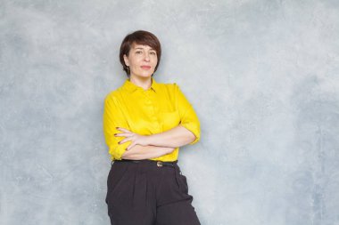Kendine güveni tam, orta yaşlı iş kadını, sarı gömlekli, gri elbiseli. 