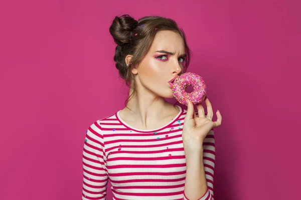 Diet concept. Doubting woman eating donut on vivid pink background