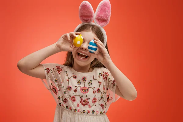 Cute little child wearing bunny ears on Easter day. Girl with painted eggs on bright red background.