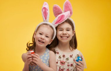 Paskalya günü tavşan kulağı takan şirin çocuklar. Parlak sarı arka planda boyalı yumurtaları olan kızlar.                               