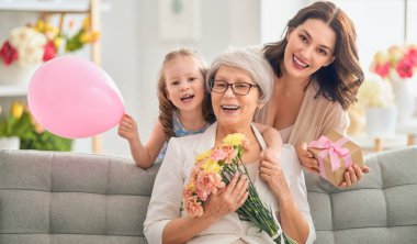 Anneler Günün kutlu olsun! Çocuk kızı, anne ve büyükannenin onlara çiçek ve hediye vermesini kutluyor. Büyükanne, anne ve kız gülümsüyor ve sarılıyorlar. Aile tatili ve birliktelik.