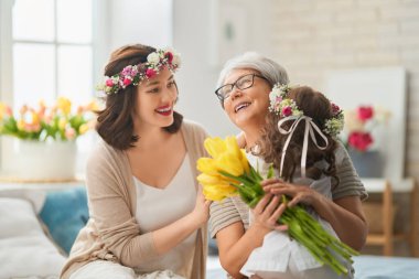 Anneler Günün kutlu olsun! Çocuk kızı, anne ve büyükannenin onlara çiçek ve hediye vermesini kutluyor. Büyükanne, anne ve kız gülümsüyor ve sarılıyorlar. Aile tatili ve birliktelik.