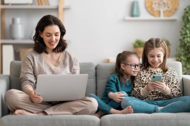 Sevgi dolu bir aile. Laptop ve akıllı telefon kullanan genç anne ve kız çocukları. Komik anne ve güzel çocuklar evde kalarak eğleniyorlar.. 