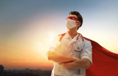 Koronavirüs salgını sırasında maskeli ve süper kahraman pelerinli bir doktor. Virüs ve hastalık koruması, karantina. COVID-2019. Tıbbın süper kahraman gücü. Günbatımı şehir planındaki kişi.