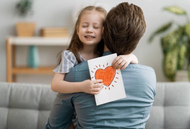 Babalar günün kutlu olsun! Çocuk kızı babasını tebrik ediyor ve ona kartpostal veriyor. Baba ve kız gülümsüyor ve sarılıyorlar. Aile tatili ve birliktelik.