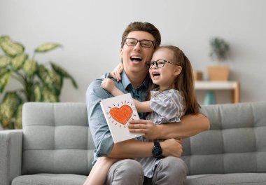 Babalar günün kutlu olsun! Çocuk kızı babasını tebrik ediyor ve ona kartpostal veriyor. Baba ve kız gülümsüyor ve sarılıyorlar. Aile tatili ve birliktelik.