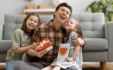 Babalar günün kutlu olsun! Çocukların kızları babalarını tebrik ediyor ve ona kartpostal veriyorlar. Baba ve kızlar gülümsüyor ve sarılıyorlar. Aile tatili ve birliktelik.