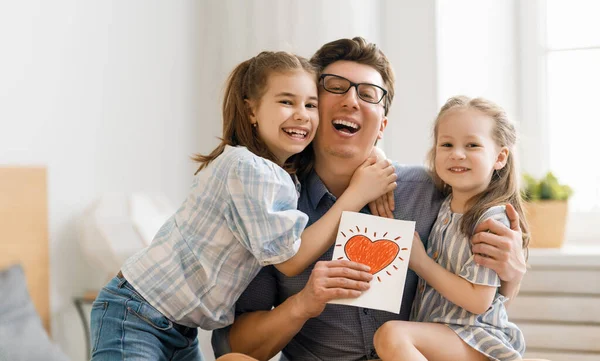 Babalar günün kutlu olsun! Çocukların kızları babalarını tebrik ediyor ve ona kartpostal veriyorlar. Baba ve kızlar gülümsüyor ve sarılıyorlar. Aile tatili ve birliktelik.