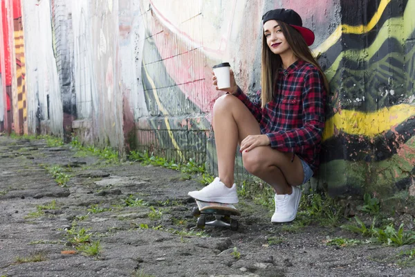 Chica joven de moda sentada con un café y un monopatín — Foto de Stock