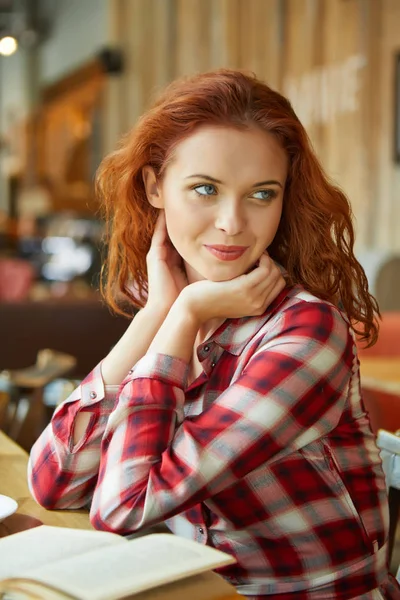 मुस्कुराते लाल बालों वाली महिला कॉफी की दुकान पर किताब पढ़ रही है — स्टॉक फ़ोटो, इमेज