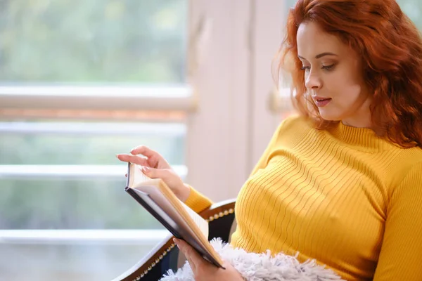 Genç kızıl saçlı kadın pencere kenarında otururken kitap okuyor. — Stok fotoğraf