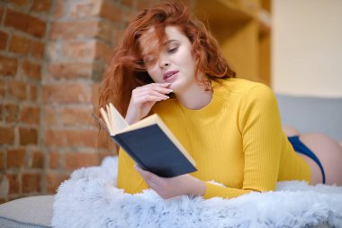 Kızıl saçlı kadın evde kitap okuyor.