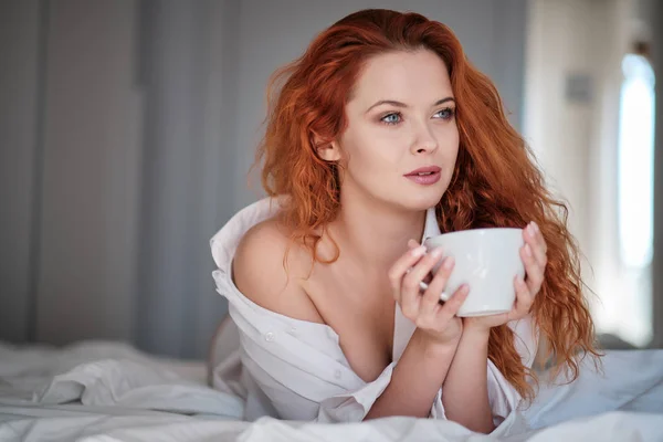 Aantrekkelijke roodharige vrouw met een kopje koffie op het bed — Stockfoto