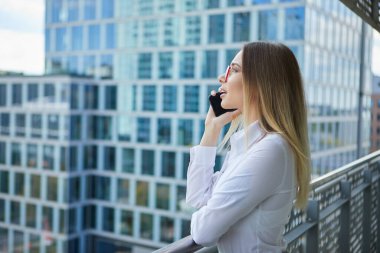 Emlakçı telefonda konuşuyor. Arka planda modern binalar, ofis binaları.