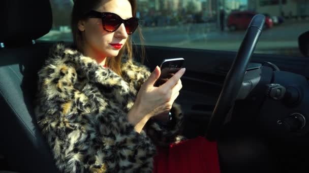 美丽的女人，一件裘皮大衣，使用智能手机，在车里喝咖啡 — 图库视频影像