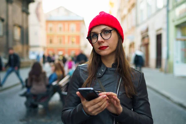 Γυναίκα με κόκκινα χείλη και κόκκινο καπέλο χρησιμοποιεί smartphone και βόλτες alon — Φωτογραφία Αρχείου