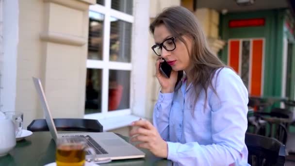 Hermosa joven mujer trabajando y hablando en un teléfono inteligente en un café al aire libre — Vídeos de Stock