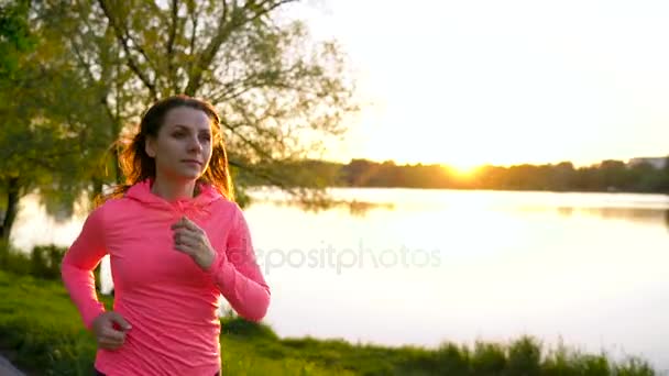 La mujer corre por el parque en la orilla del lago al atardecer, en cámara lenta — Vídeo de stock