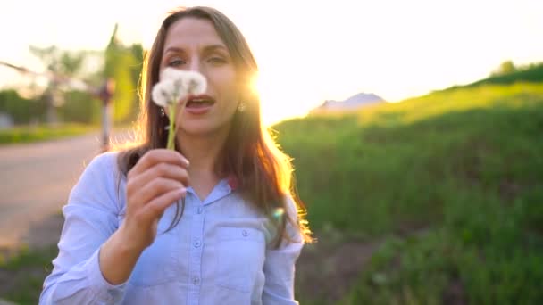 Belleza mujer soplando diente de león contra el atardecer, cámara lenta — Vídeo de stock