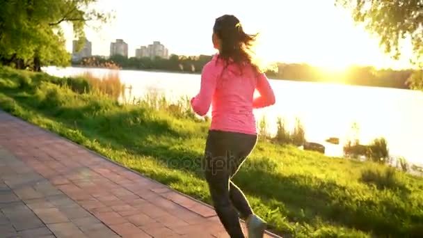 La mujer corre por el parque en la orilla del lago al atardecer, en cámara lenta — Vídeos de Stock