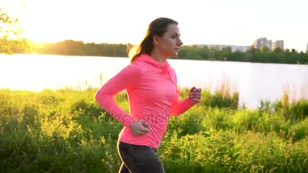 Mulher corre através do parque na costa do lago ao pôr do sol, câmera lenta — Vídeo de Stock