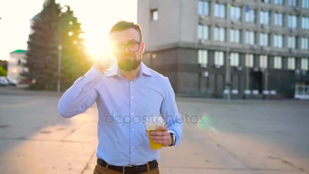 スマート フォンで話していると、通りを歩きながらジュースを飲む男します。 — ストック動画