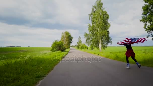 Young teenage woman waving a US flag is roller skating on the rural road — Stock Video