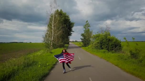 Молодая девушка-подросток, размахивающая флагом США, катается на роликах по сельской дороге — стоковое видео